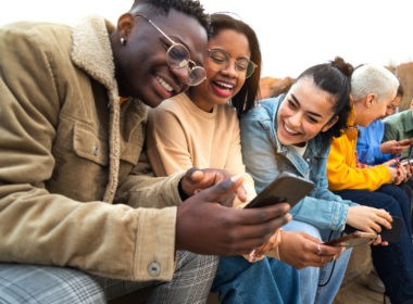 Happy multiracial teen college students having fun using mobile phone together in campus outdoors.