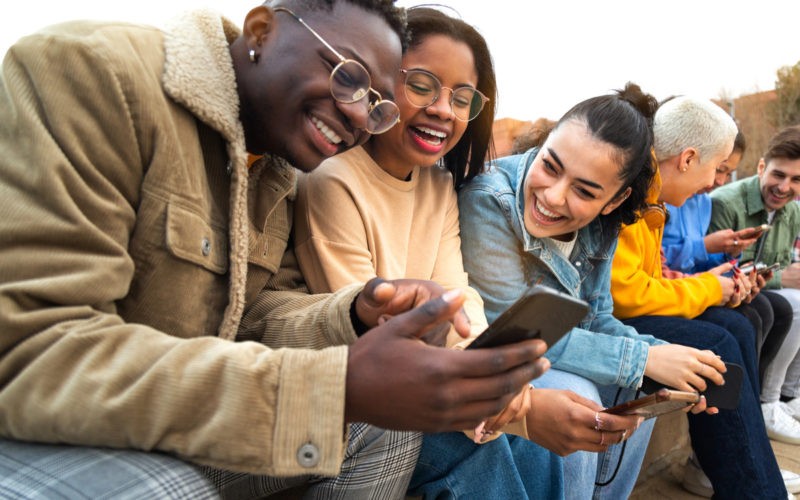 Happy multiracial teen college students having fun using mobile phone together in campus outdoors.