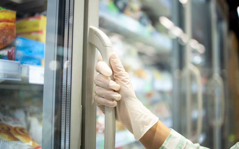 Close up view of hand in protective rubber gloves opening fridge with frozen food in supermarket