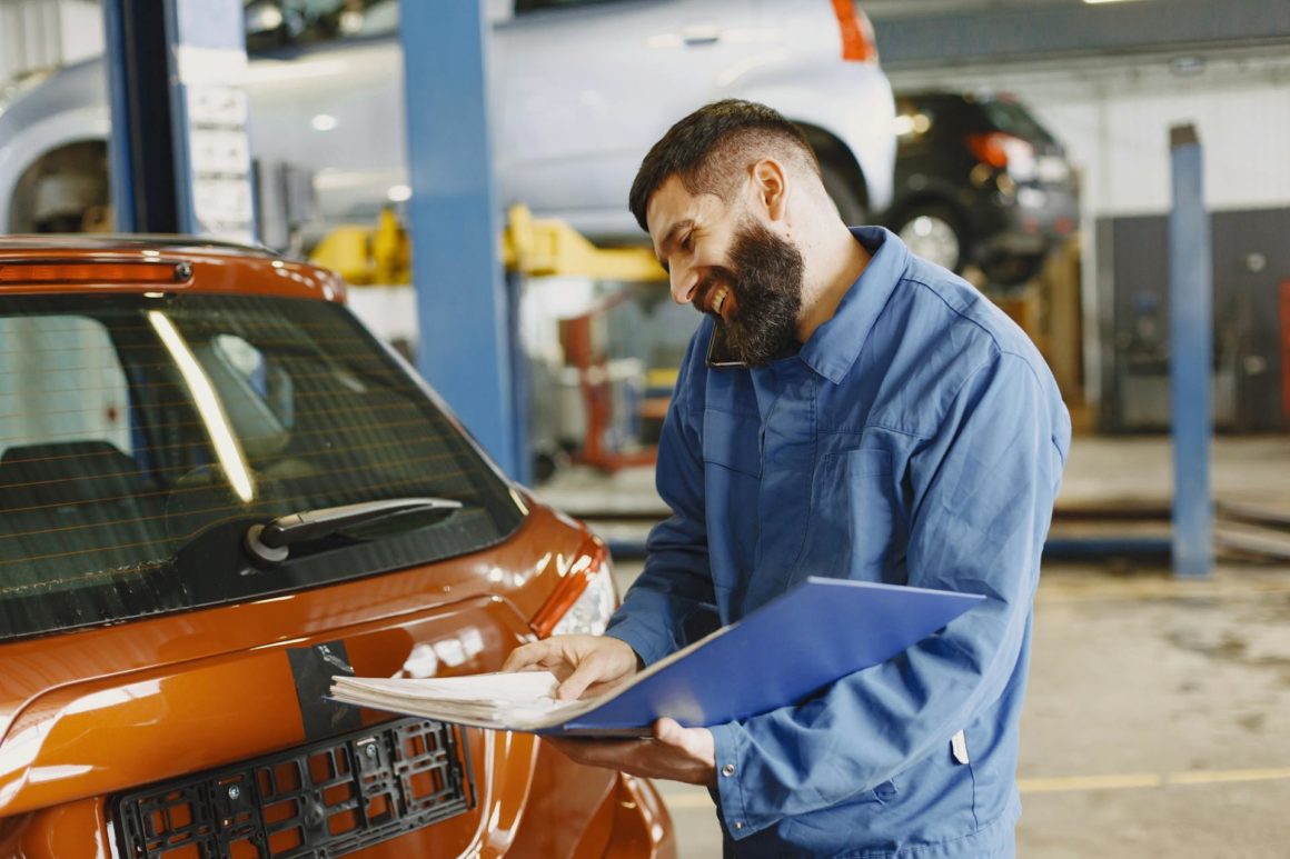 Smiling Man in Blue Coveralls Standing Beside an Orange Car Having Conversation Over the Phone