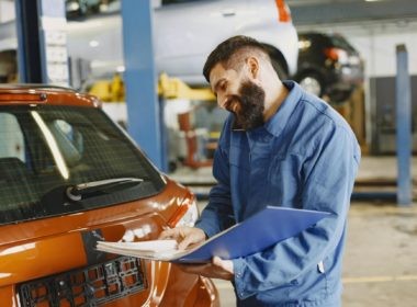 Smiling Man in Blue Coveralls Standing Beside an Orange Car Having Conversation Over the Phone