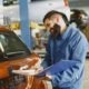 Smiling Man in Blue Coveralls Standing Beside an Orange Car Having Conversation Over the Phone
