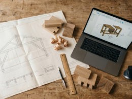 Overhead view of a woodworking workspace with CAD drawings, a laptop and wood samples on a clean desk