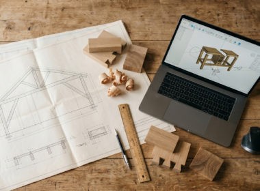 Overhead view of a woodworking workspace with CAD drawings, a laptop and wood samples on a clean desk