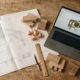 Overhead view of a woodworking workspace with CAD drawings, a laptop and wood samples on a clean desk
